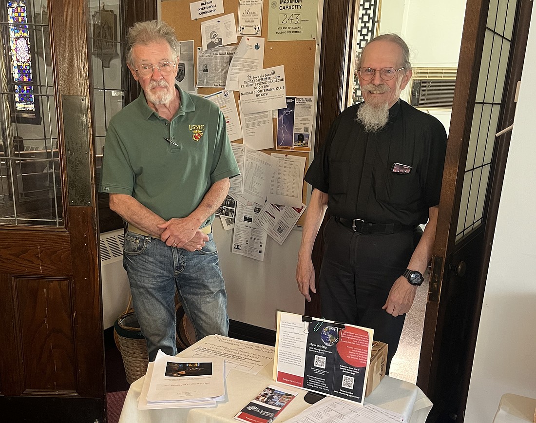 Ken Scallon, parishioner of St. Mary’s of the Nativity, and Father John Close, sacramental minister, stand next to a station in the front entrance of the church that has information about the refugee program the church donates to and a spot for donations to be dropped off. (Emily Benson photos)